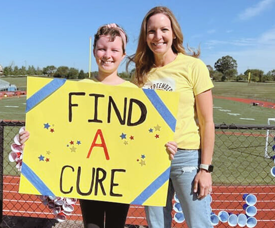 Rylee and Crystal smiling outdoors holding a sign that reads "find a cure"